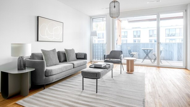 A modern living room with a grey sofa, a coffee table, a side table with a lamp, and a large window with a view of buildings. - Lux Wollishofen 2.5 Room w/ W/D & Storage, 2 min to Market, by Blueground (Zurich)