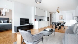 A modern living room with a sofa, a coffee table, and a TV. There is a kitchen area with cabinets and a dining table. - Lux Wollishofen 2.5 Room w/ W/D & Storage, 2 min to Market, by Blueground (Zurich)