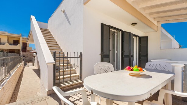 A white outdoor patio with a round table and chairs, a bowl of fruit, and a staircase leading to an upper level.
