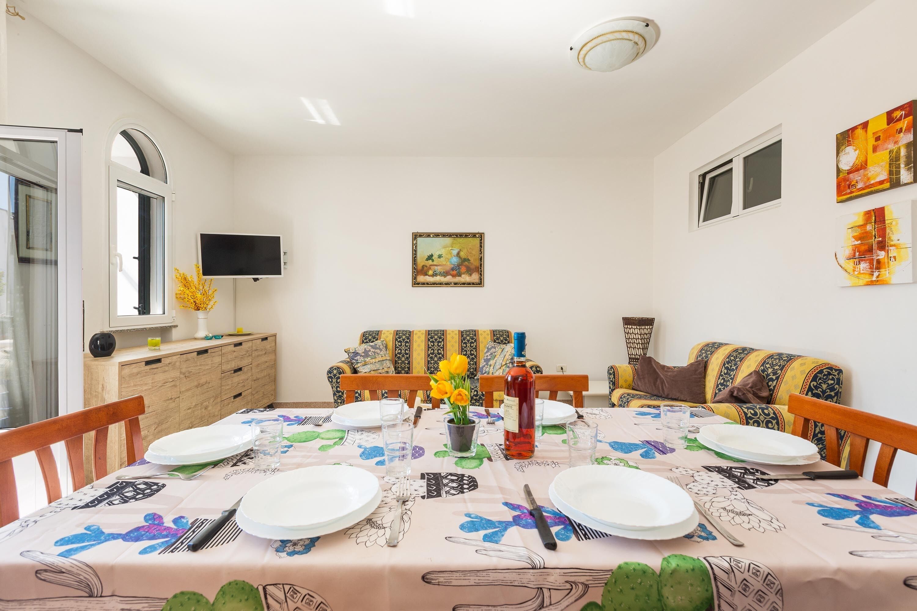 A dining area with a round table set for four, a television mounted on the wall, and a sofa in the background.