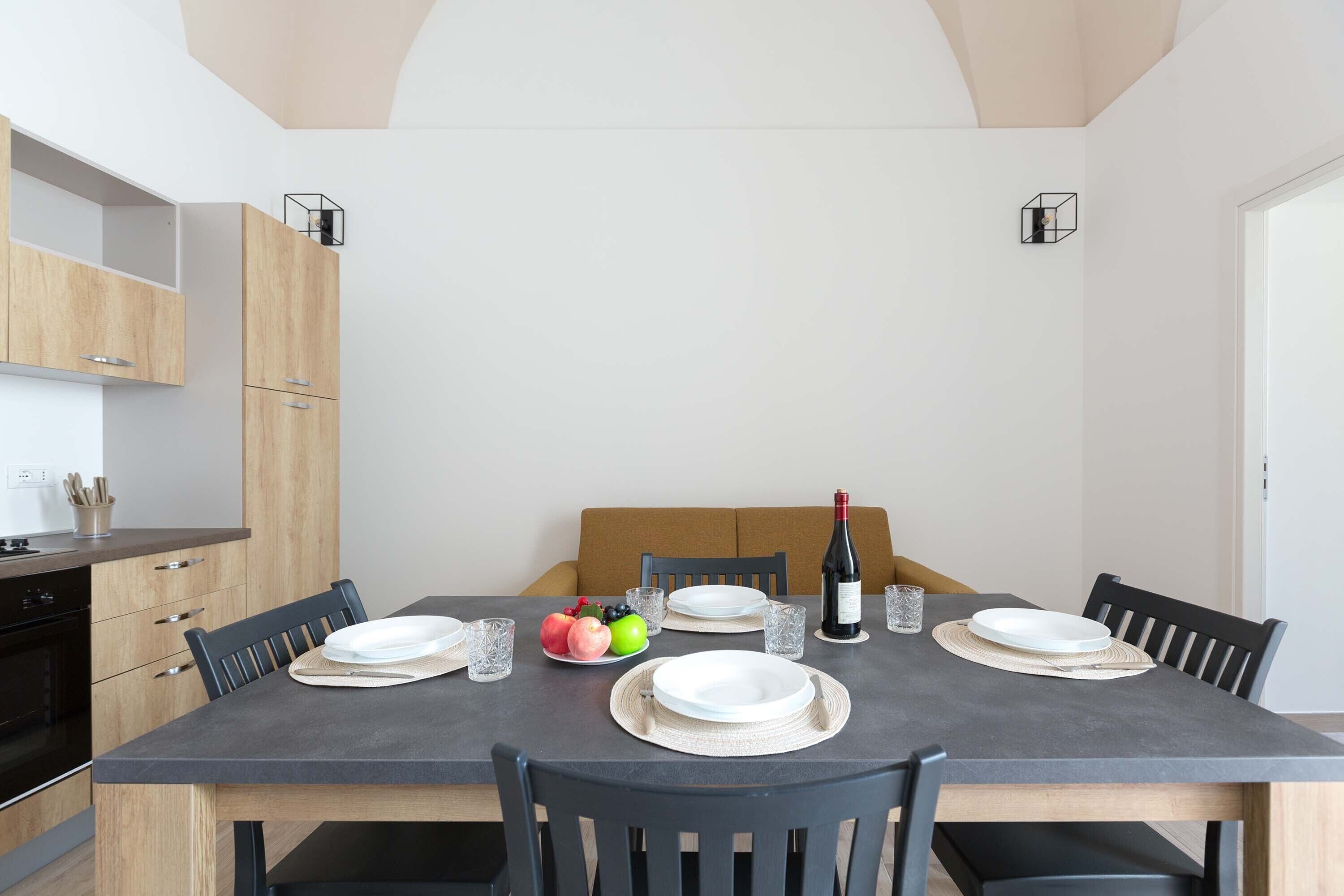 Un comedor con una mesa oscura dispuesta para cuatro personas, rodeado de armarios de madera y un sofá.