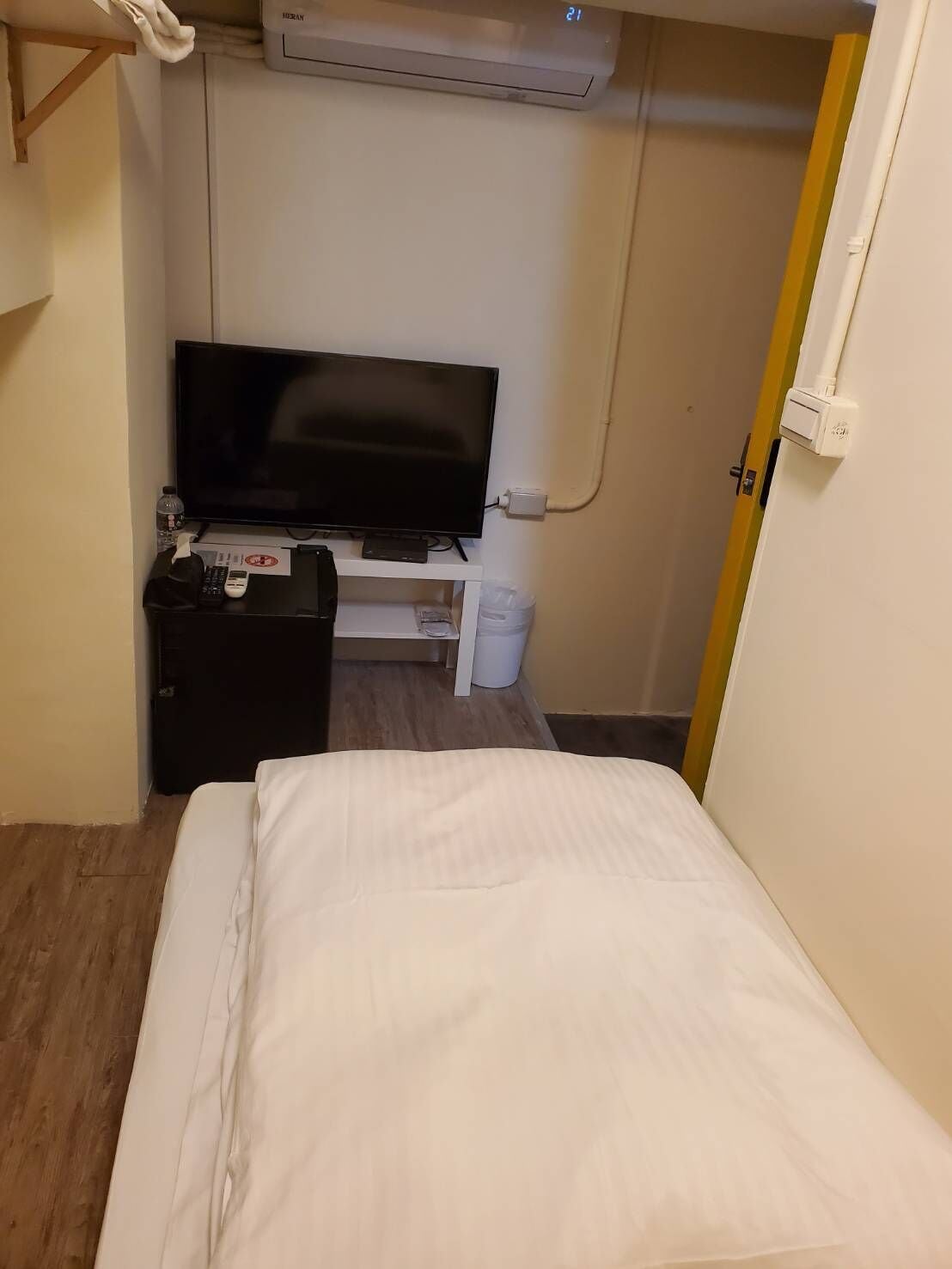 A hotel room with a bed, a television on a stand, a small table with a remote control, a water bottle, and a trash bin.