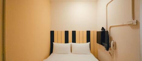 A narrow hotel room with a bed, a wall-mounted TV, and a ceiling light.