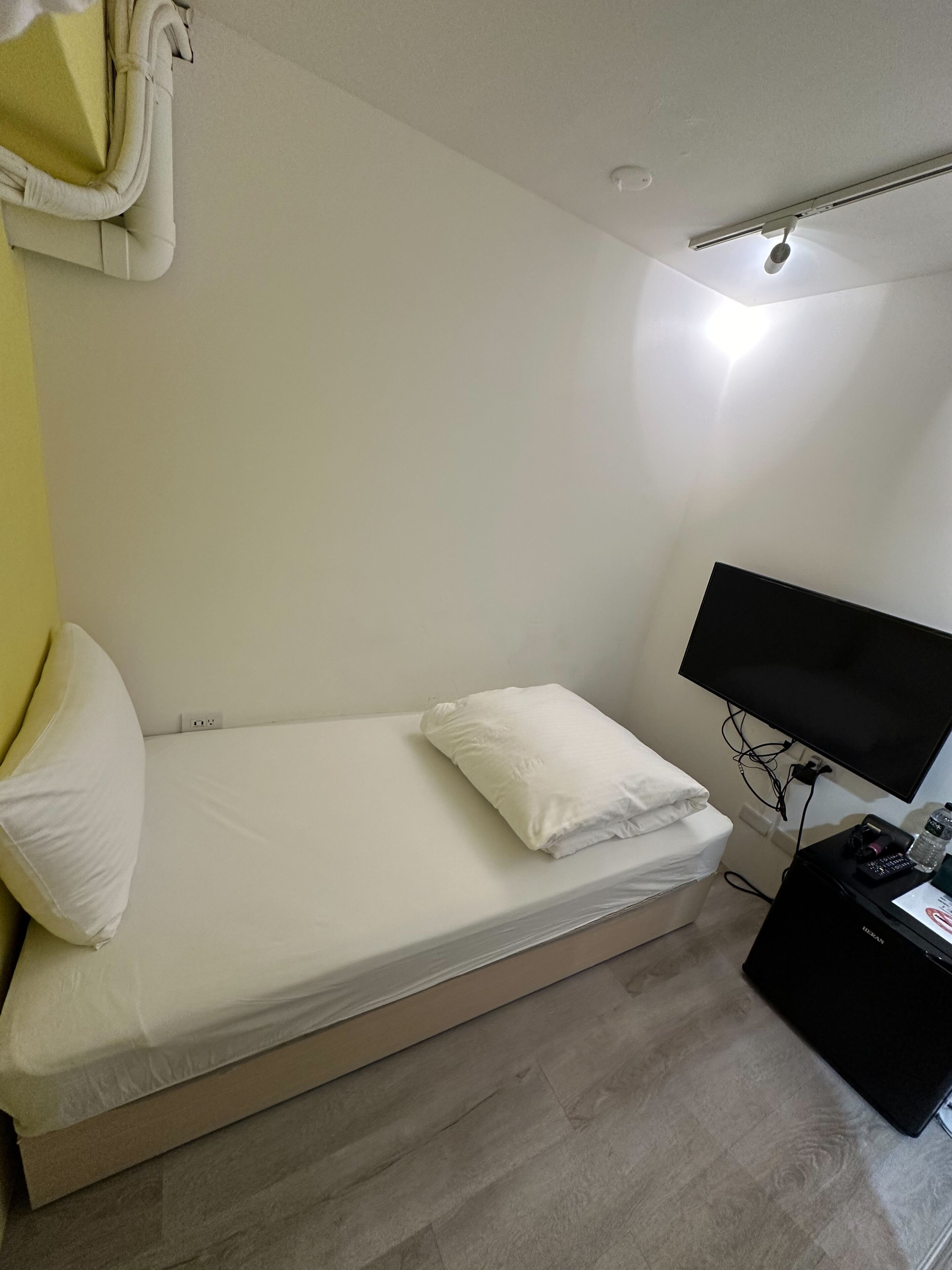 Una cama pequeña y bien arreglada con sábanas y almohadas blancas, un televisor de pantalla plana en la pared y un dispositivo electrónico negro en el suelo.
