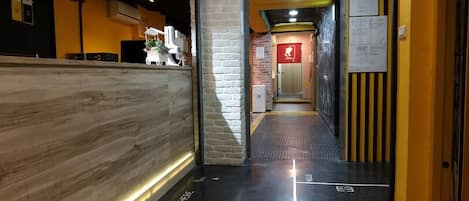 Zona de recepción de un hotel con un mostrador de madera, un pilar de ladrillo y una pared amarilla con un letrero rojo.
