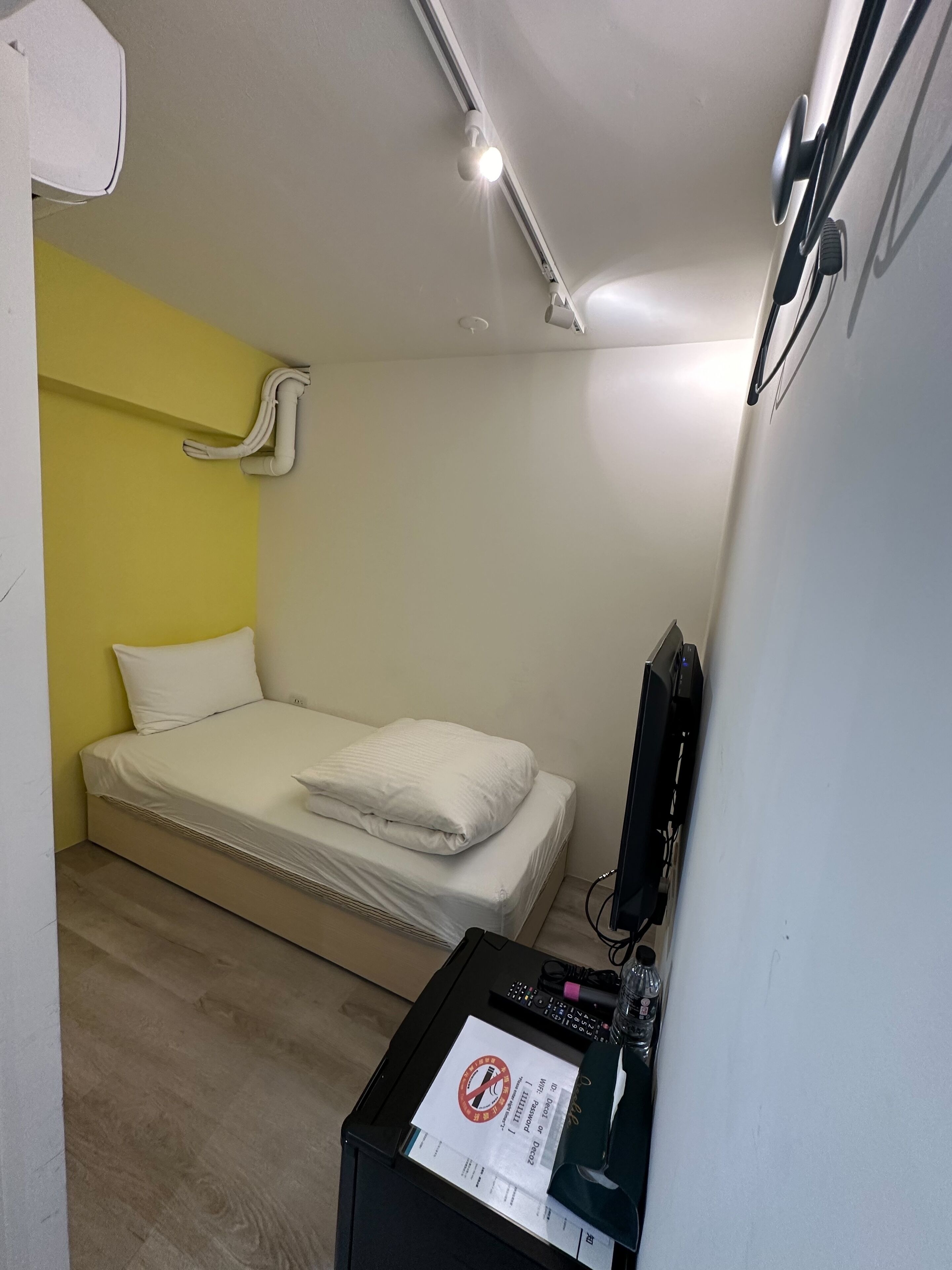 A small, neatly arranged room with a bed, a desk, and a wall-mounted TV.