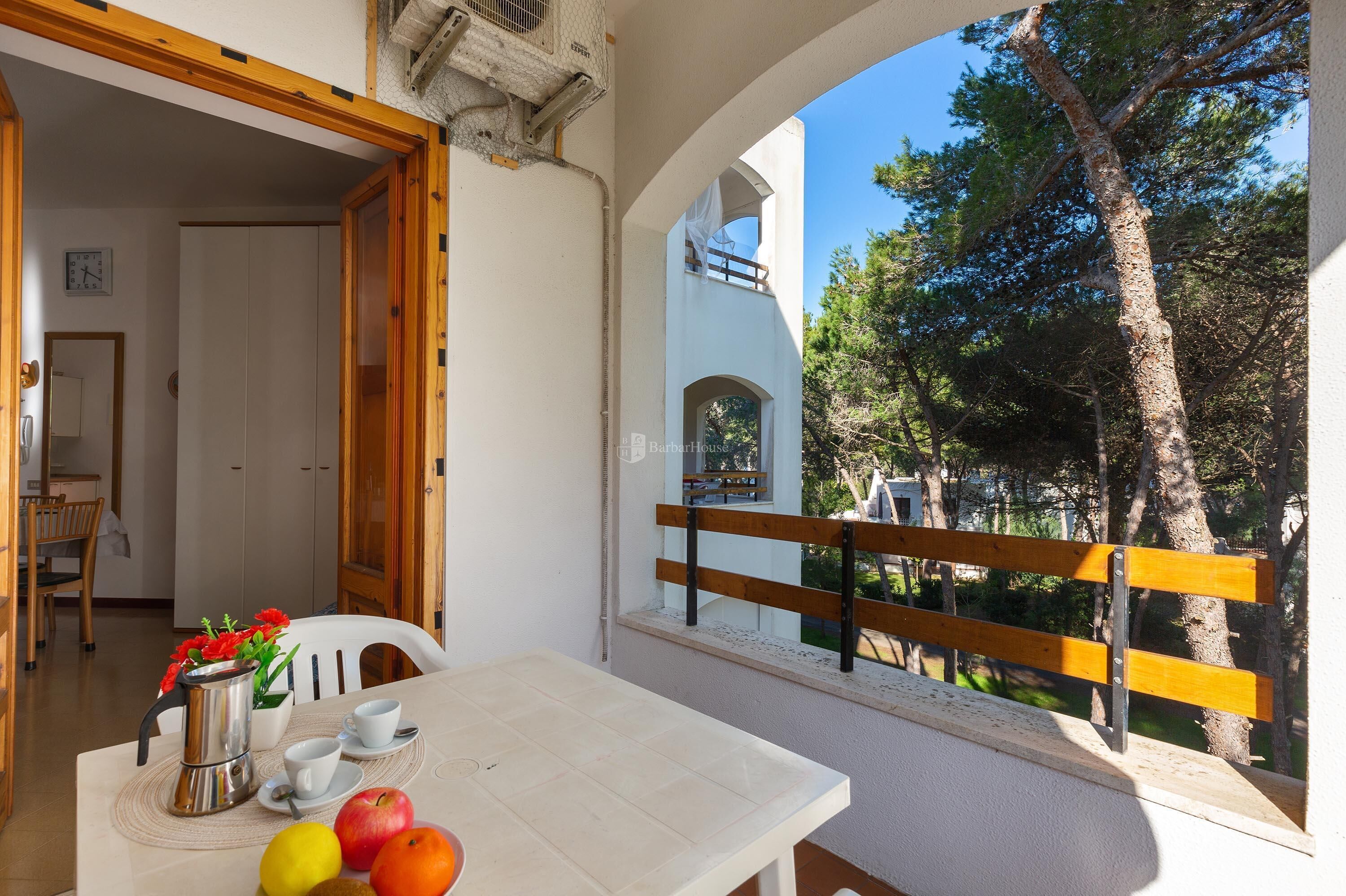 Un balcó amb una barana de fusta, una taula blanca amb fruites i una cafetera, i vista a arbres i un edifici.