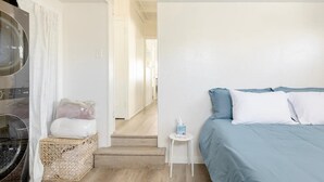 A bedroom with a bed, a small bedside table, a wicker basket, and a view of a hallway.