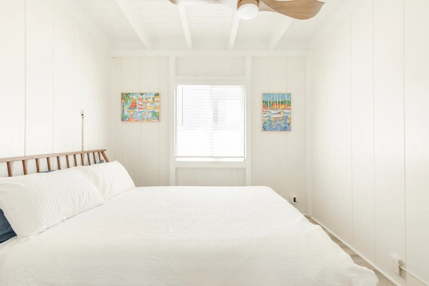 A bedroom with a bed, white walls, wooden headboard, two paintings, and a window with blinds.