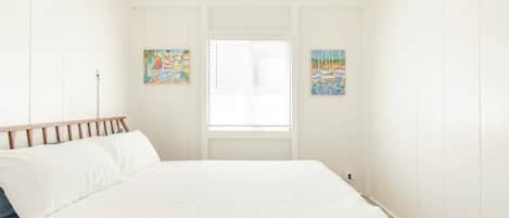 A bedroom with a bed, white walls, wooden headboard, two paintings, and a window with blinds.
