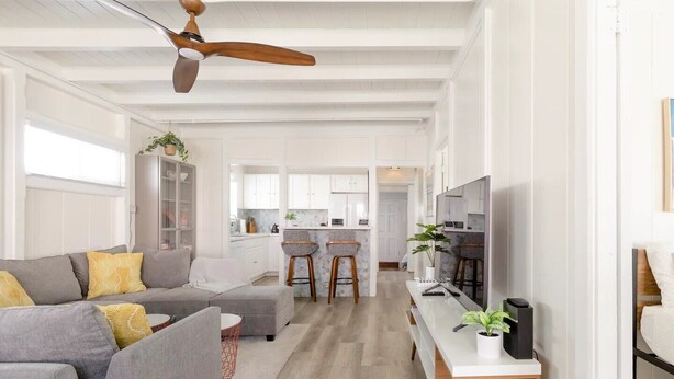 A modern living room with a grey sofa, a wooden ceiling fan, and a kitchen area in the background.