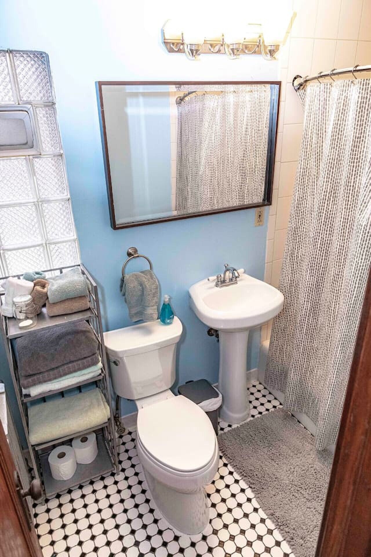 Een badkamer met toilet, wastafel en spiegel. Er is een handdoekrek met handdoeken en een fles zeep. De vloer heeft een zwart-wit geruit patroon.