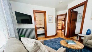 Un salon avec un canapé, une table basse et un téléviseur fixé au mur. On y trouve également un radiateur, une plante et une porte qui donne sur une autre pièce.
