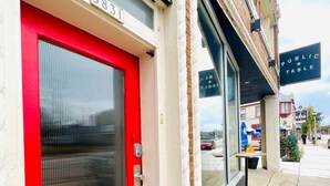 Une porte rouge avec une fenêtre, une enseigne indiquant « Table ouverte au public », et la façade d’un bâtiment en briques.