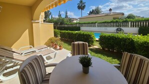 An outdoor patio with a round table and striped chairs, a small potted plant, and a view of a swimming pool and a house in the background. - Townhouse ALDEA DEL MAR with direct access to the swimming pool (Poblets (els))