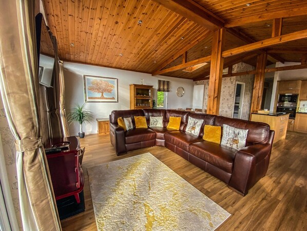 A spacious living room with a large brown leather sectional sofa, wooden ceiling beams, and a kitchen area in the background.