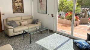 En stue med en beige sofa, et glasbord og udsigt til en have gennem en skydedør.