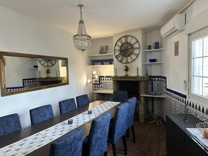 Un espace repas avec une longue table, des chaises à motifs bleus, un lustre, une grande horloge murale et une cheminée ornée de carreaux à motifs.