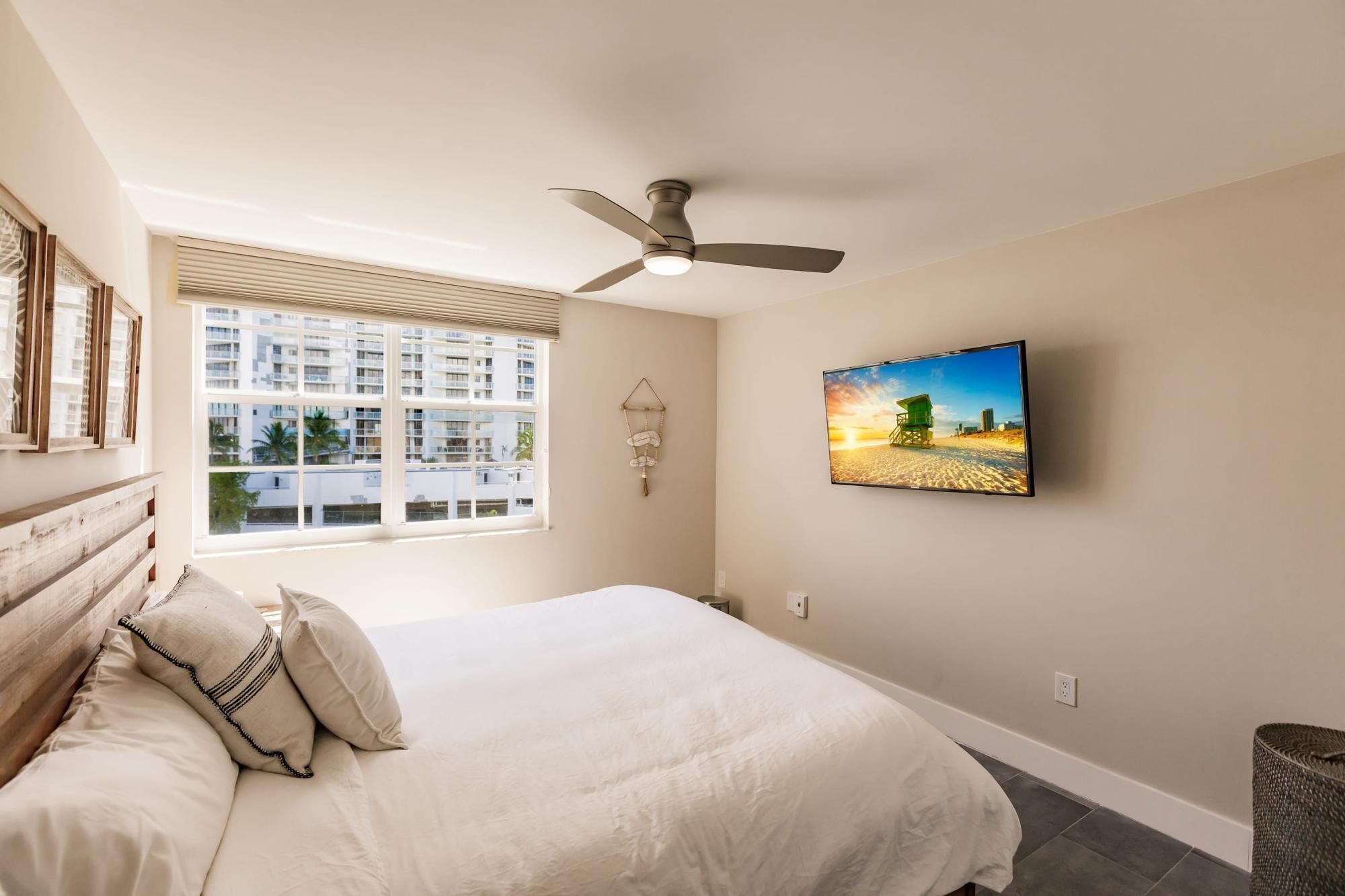 A bedroom with a bed, a ceiling fan, a large window with blinds, a wall-mounted TV, and a beach scene painting.