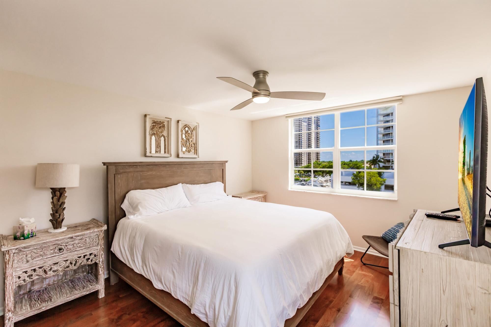 A bedroom with a bed, a ceiling fan, a nightstand, a lamp, a desk with a chair, and a large window with a view of buildings and trees.