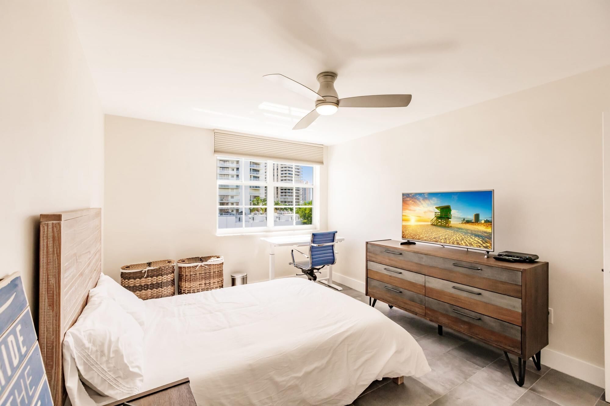 Une chambre à coucher comprenant un lit, une commode, un téléviseur, un ventilateur de plafond, une fenêtre avec des stores et une vue sur les bâtiments environnants.