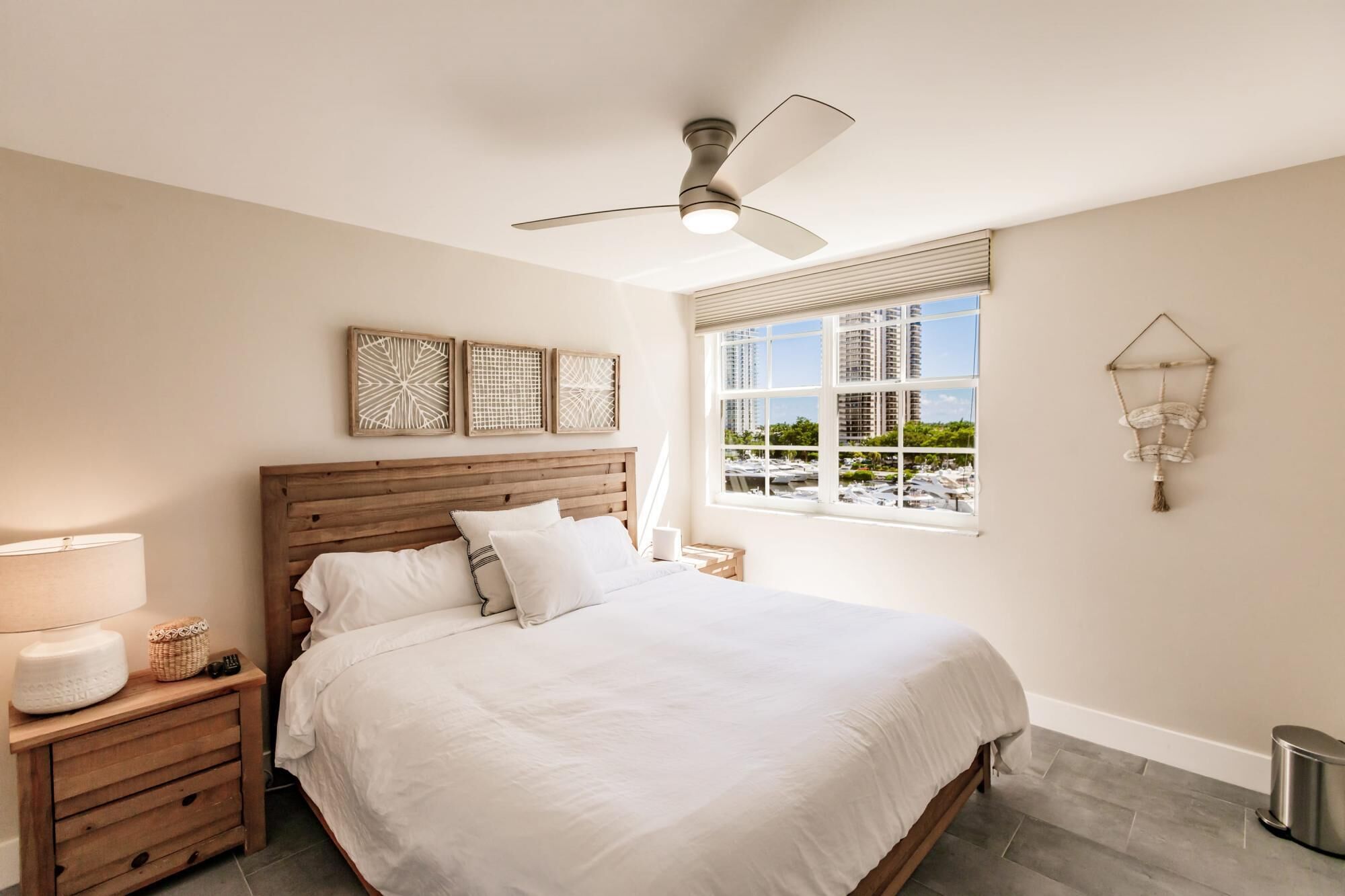 Une chambre à coucher comprenant un lit, une tête de lit en bois, des tables de chevet, un ventilateur de plafond, une fenêtre donnant sur des bâtiments et des éléments de décoration fixés au mur.
