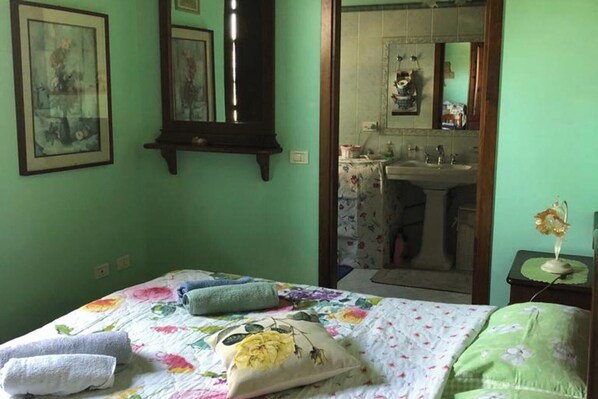 A bedroom with a floral bedspread, two pillows, a bedside table with a lamp, and a mirror above the bed.