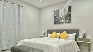 A modern bedroom with a grey tufted headboard, white bedding, and two yellow pillows. There are two bedside tables with lamps, a grey bench, and a view of the Eiffel Tower on the wall. - Modern Home In Melbourne (Melbourne)