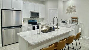 A modern kitchen with white cabinetry, stainless steel appliances, a marble countertop, and a bar area with stools. - Modern Home In Melbourne (Melbourne)