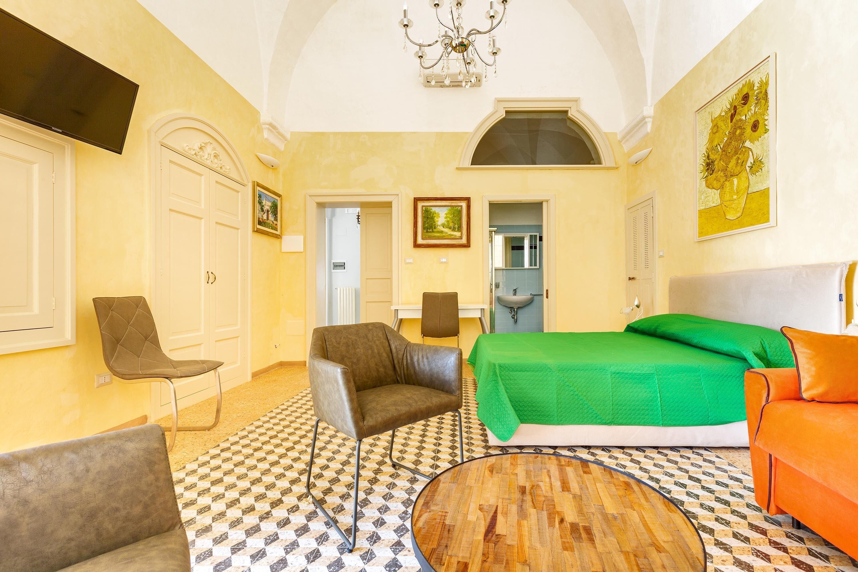 Une chambre d’hôtel avec un sol à carreaux, un lit vert, un fauteuil en cuir marron, un canapé orange, une table basse en bois et un lustre.
