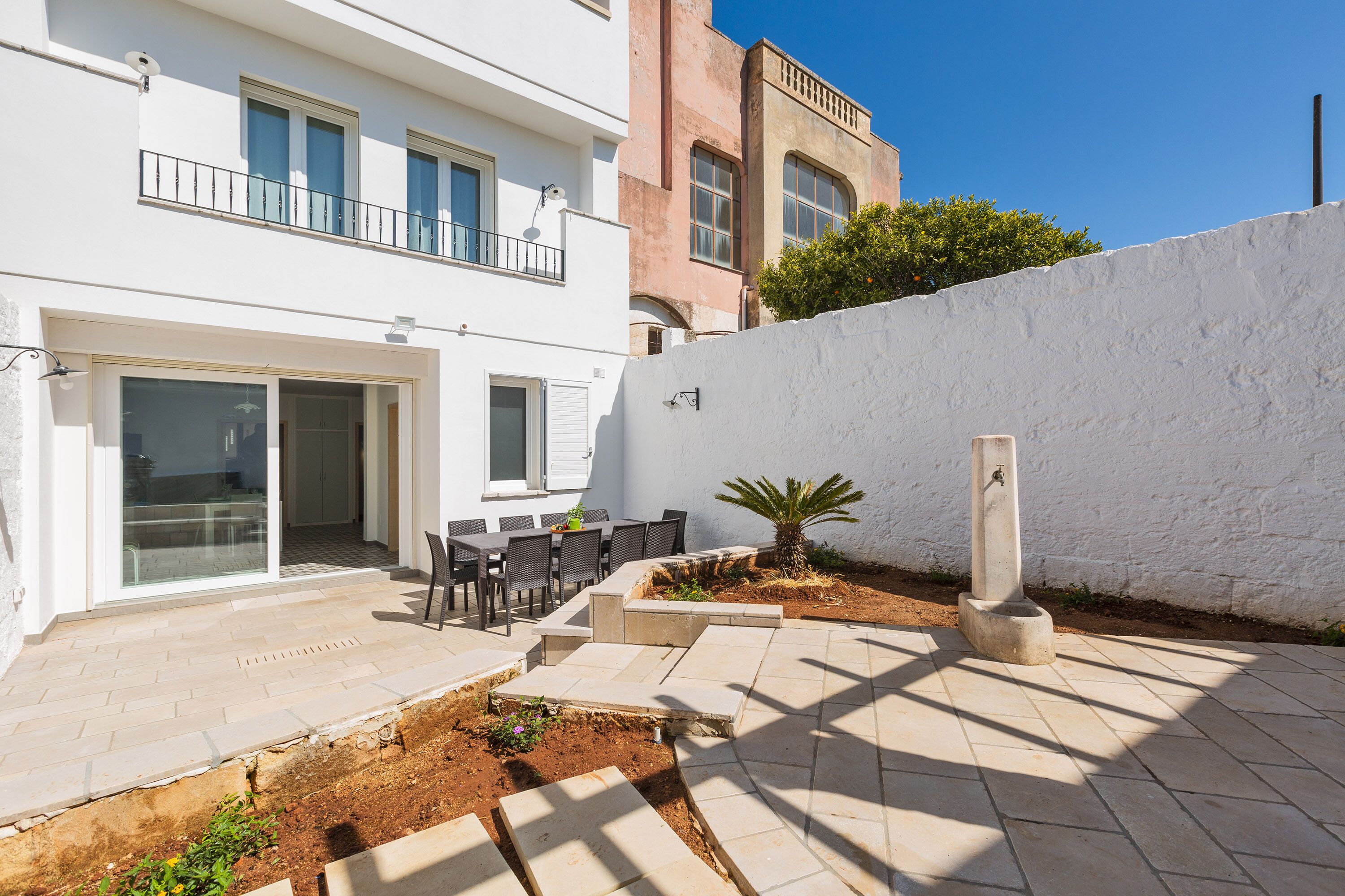 A modern building with a patio area featuring a table and chairs, a small garden, and a white wall.
