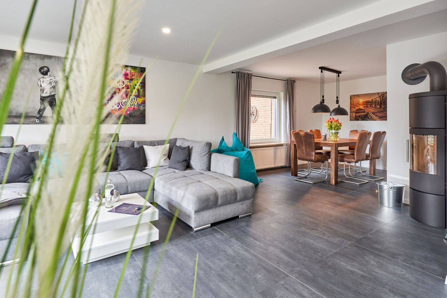 A modern living room with a grey sectional sofa, a dining area with a wooden table and chairs, and a black built-in oven.