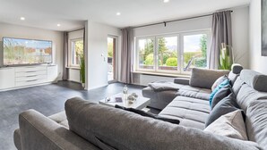 A modern living room with a grey sofa set, a flat-screen TV, and large windows offering a view of greenery outside. - Large time-out apartment - modern, close to the beach and with a large garden terrace - (Neustadt in Holstein)
