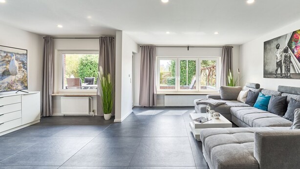 A spacious living room with a grey sofa, a coffee table, and a large window with curtains. - Large time-out apartment - modern, close to the beach and with a large garden terrace - (Neustadt in Holstein)