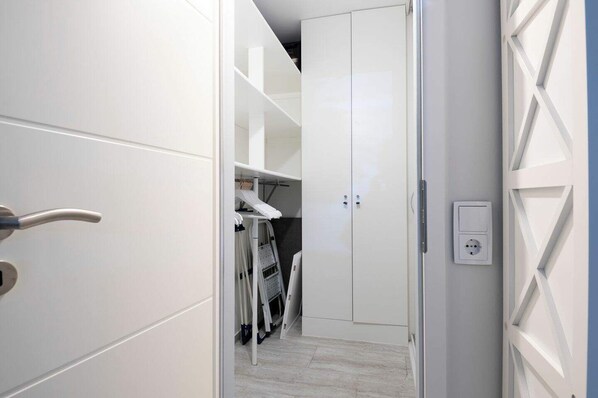 A white closet with shelves and hanging space, a white door, and a light switch on the wall.