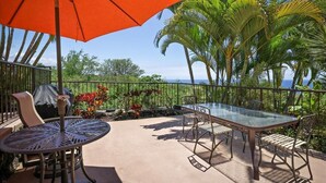 Une terrasse extérieure avec une table et des chaises en verre, un parasol rouge et une vue sur les palmiers et l’océan.