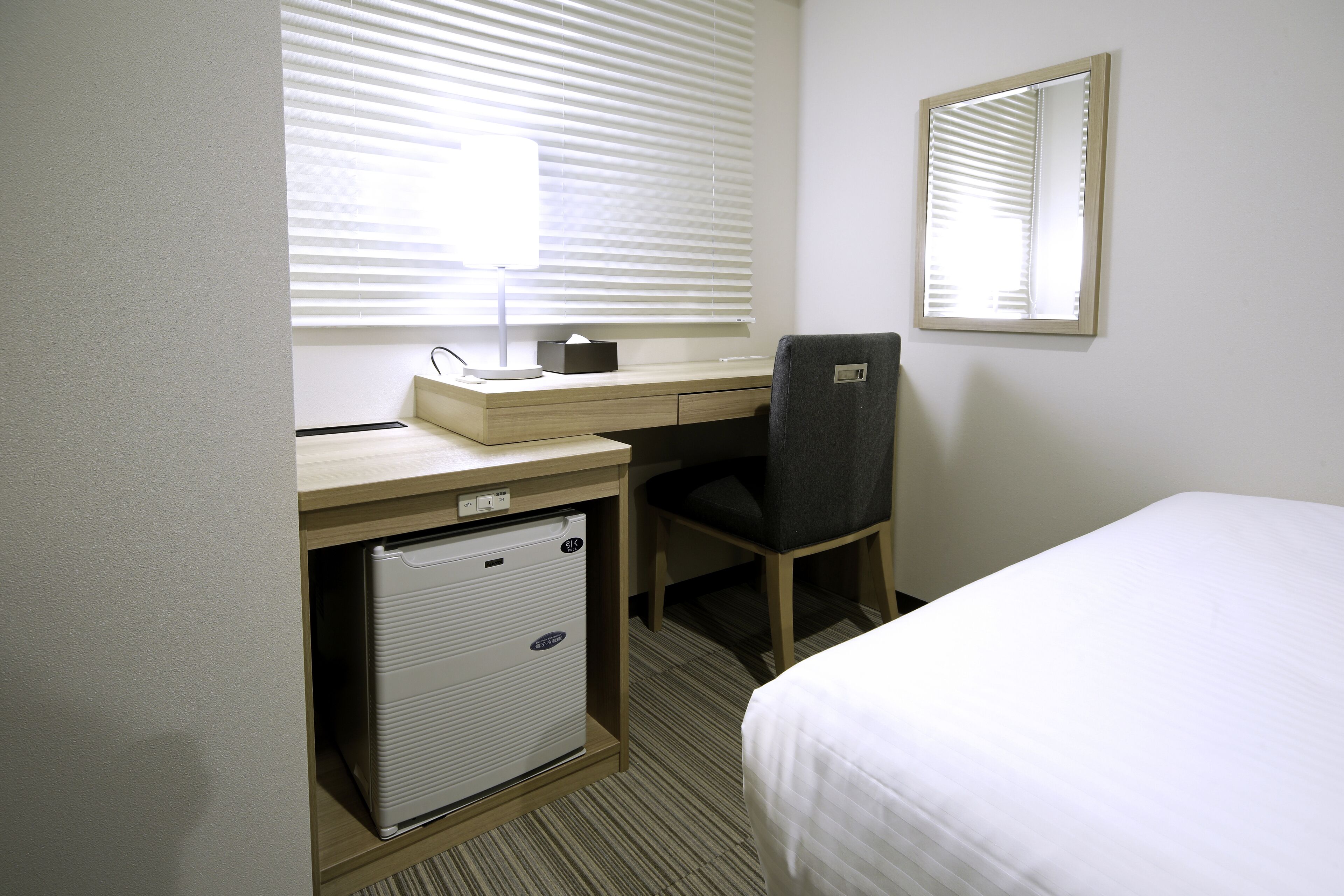 A hotel room with a bed, a desk with a computer, a chair, a small refrigerator, and a window with blinds.