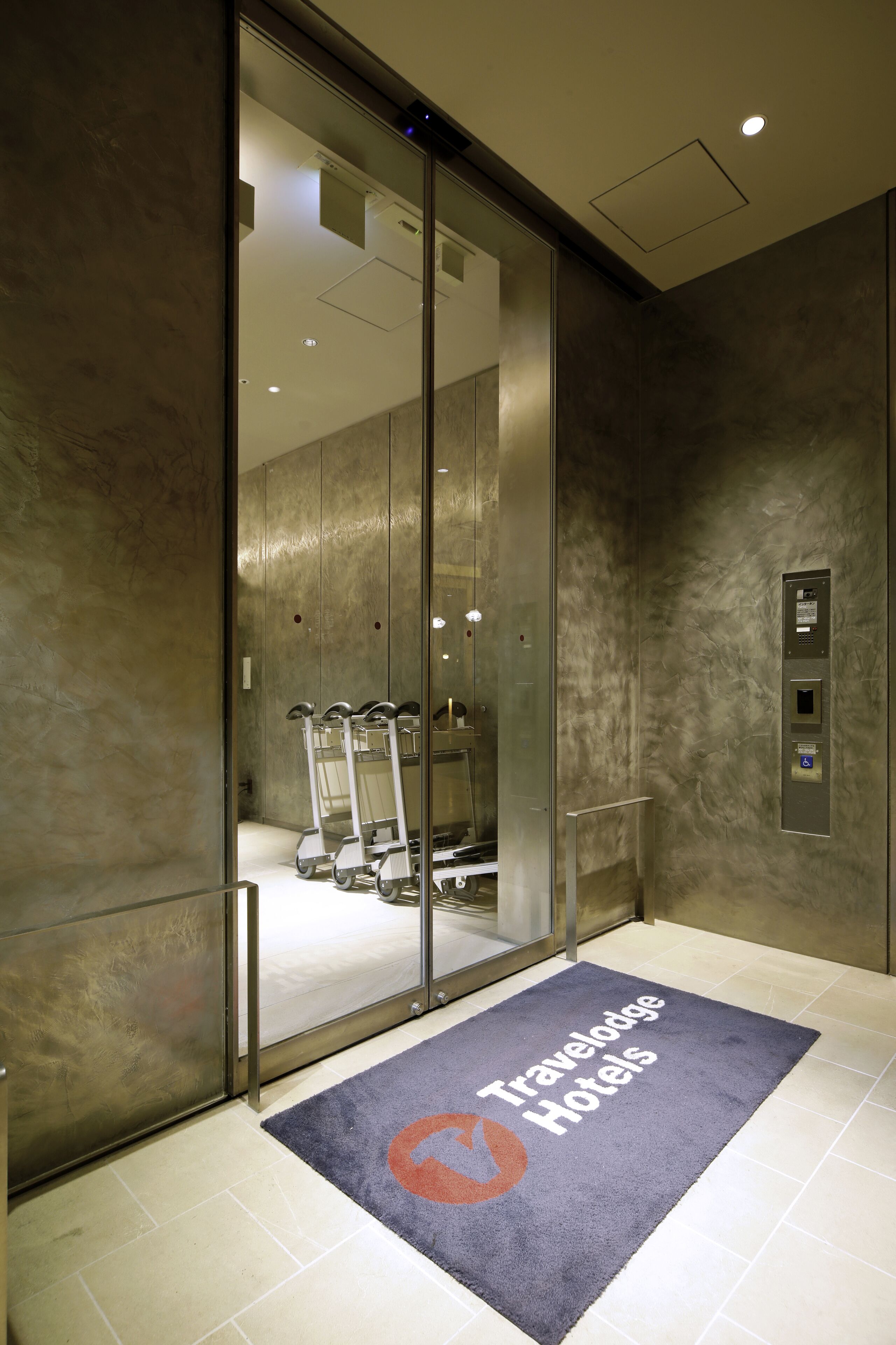 An elevator with a glass door, a luggage cart, and a mat with the Travelodge Hotels logo.
