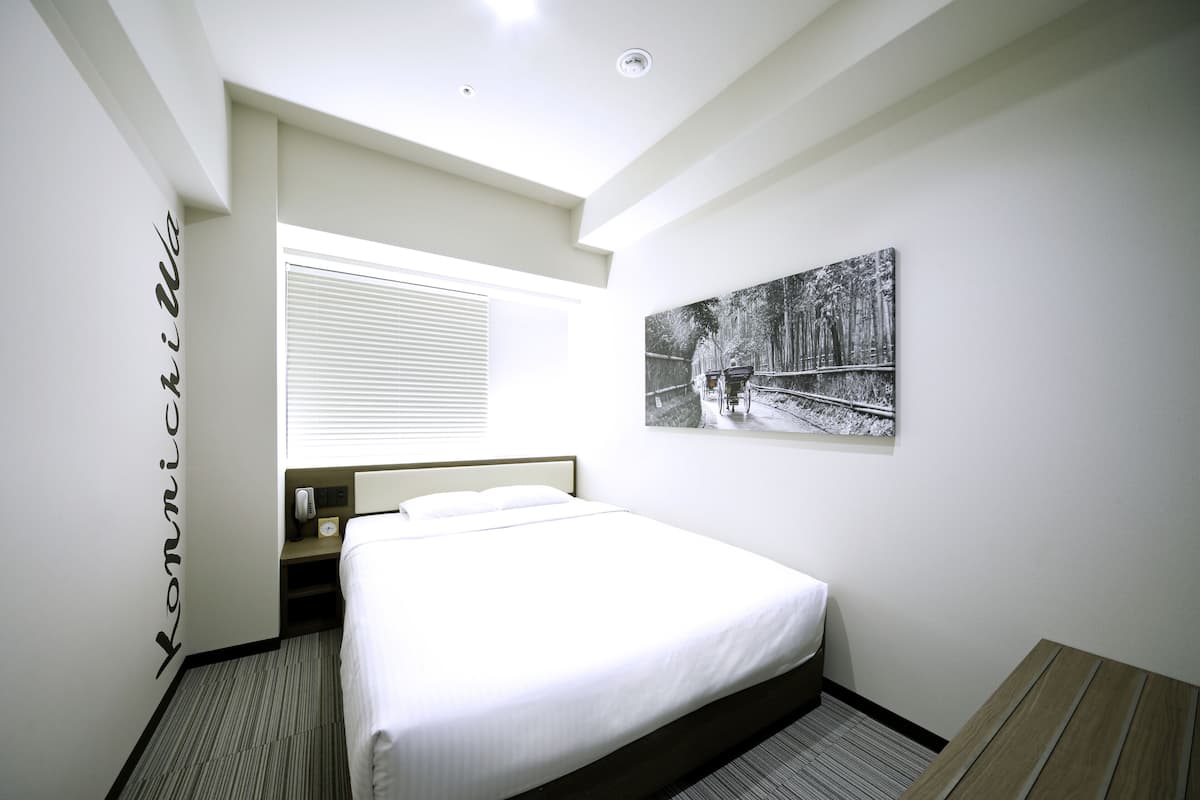 A hotel room with a bed, a bedside table, a nightstand, a window with blinds, a painting on the wall, and a sign on the wall.
