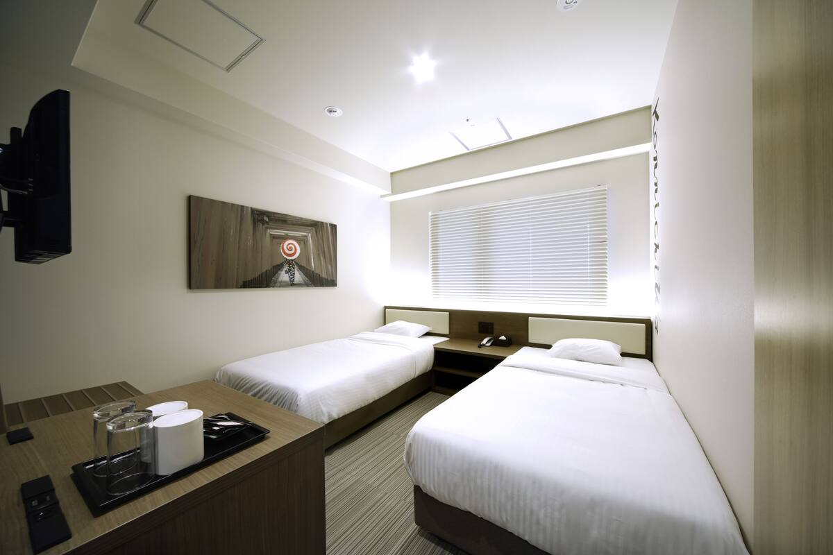 A hotel room with two single beds, a desk with a tray of amenities, a painting on the wall, and a window with blinds.