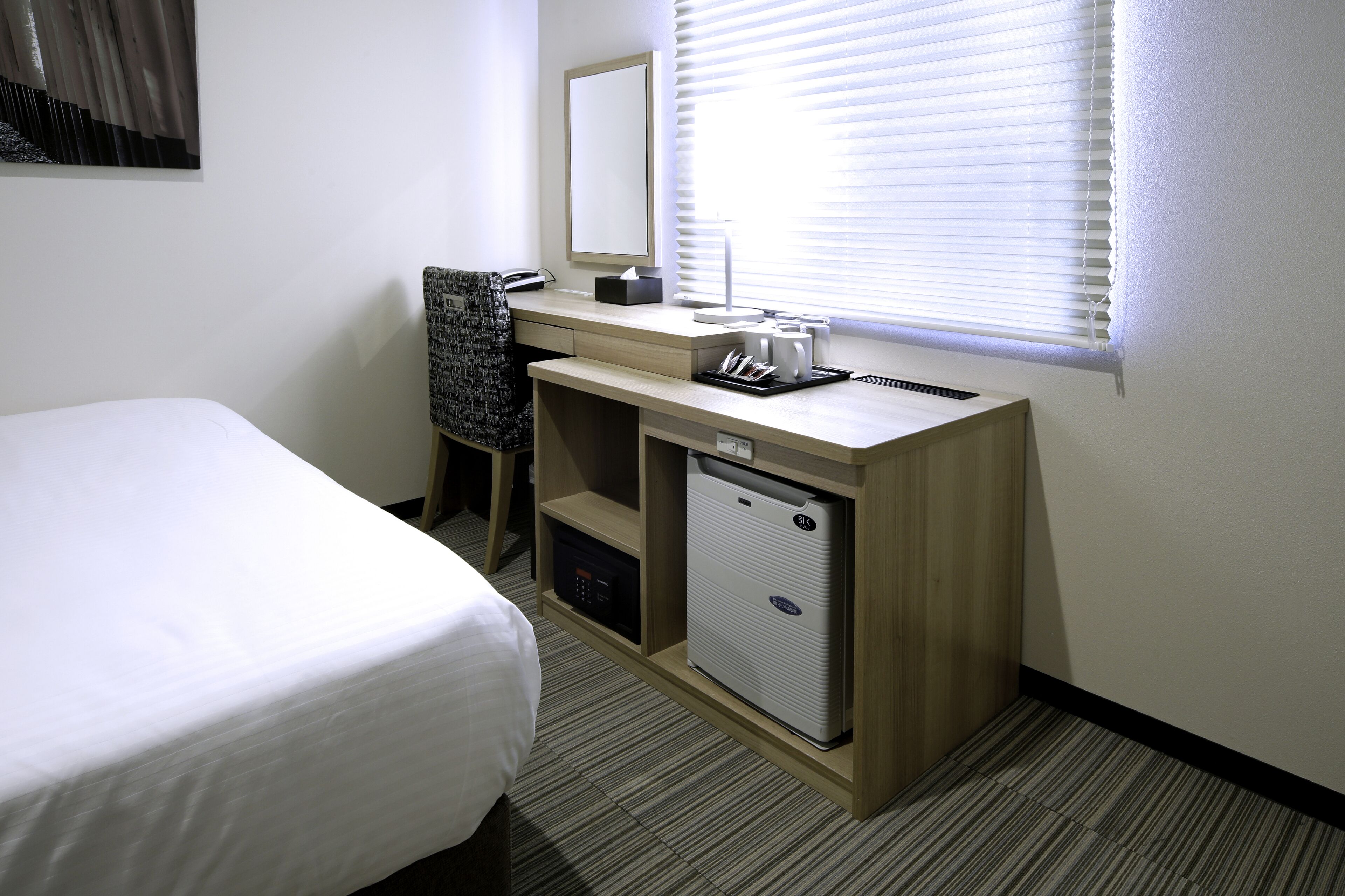 A hotel room with a bed, a desk with a computer, a small refrigerator, and a window with blinds.