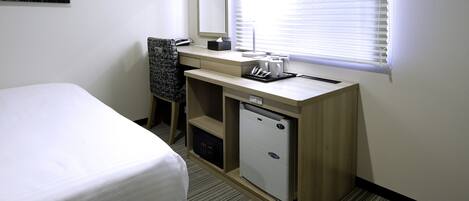 A hotel room with a bed, a desk with a computer, a small refrigerator, and a window with blinds.