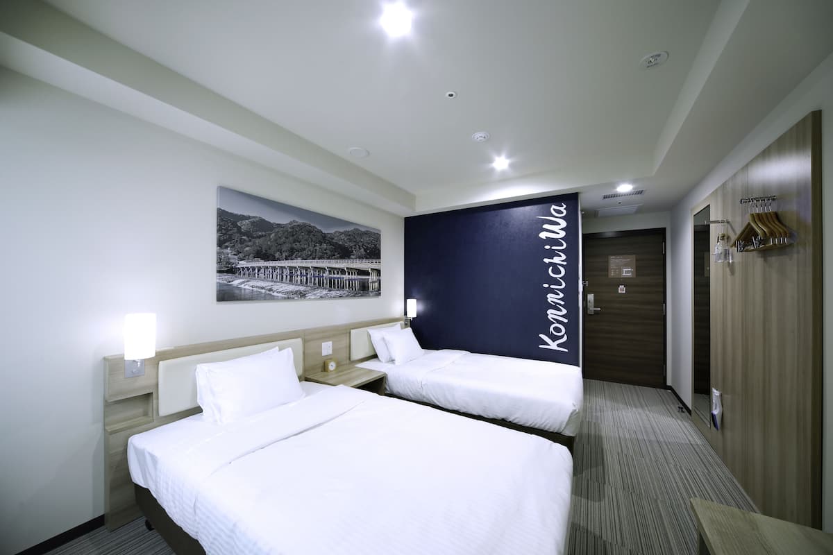 A hotel room with two beds, a nightstand, a wall-mounted lamp, a framed picture, and a dark-colored wall with the word "Kaminvilla".