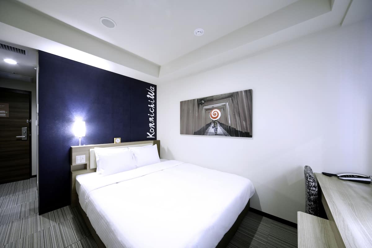 A hotel room with a bed, a desk, a chair, a nightstand, a wall-mounted lamp, and a picture of a spiral staircase.