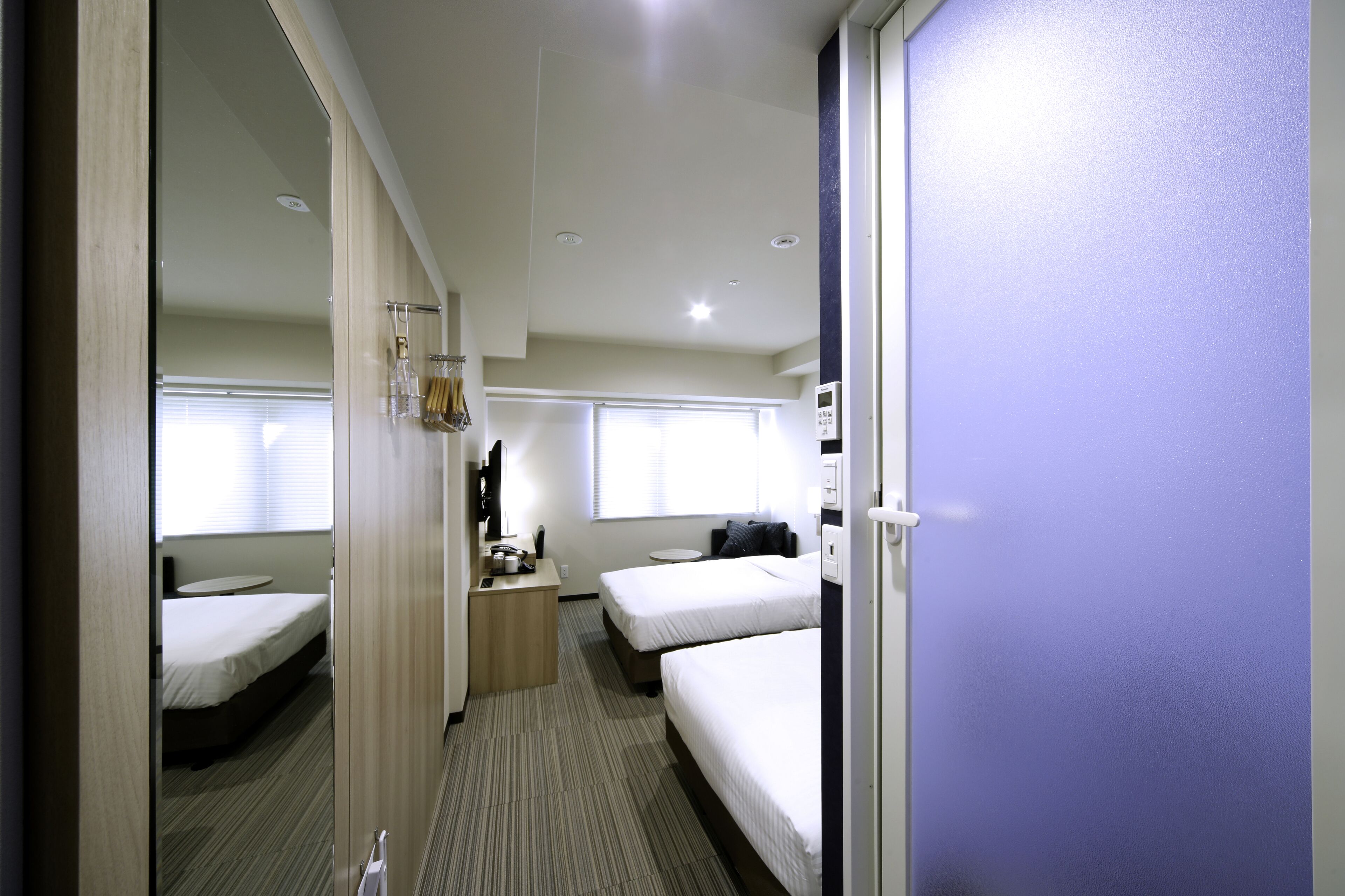 A hotel room with two single beds, a desk with a chair, a small table, and a mirror.