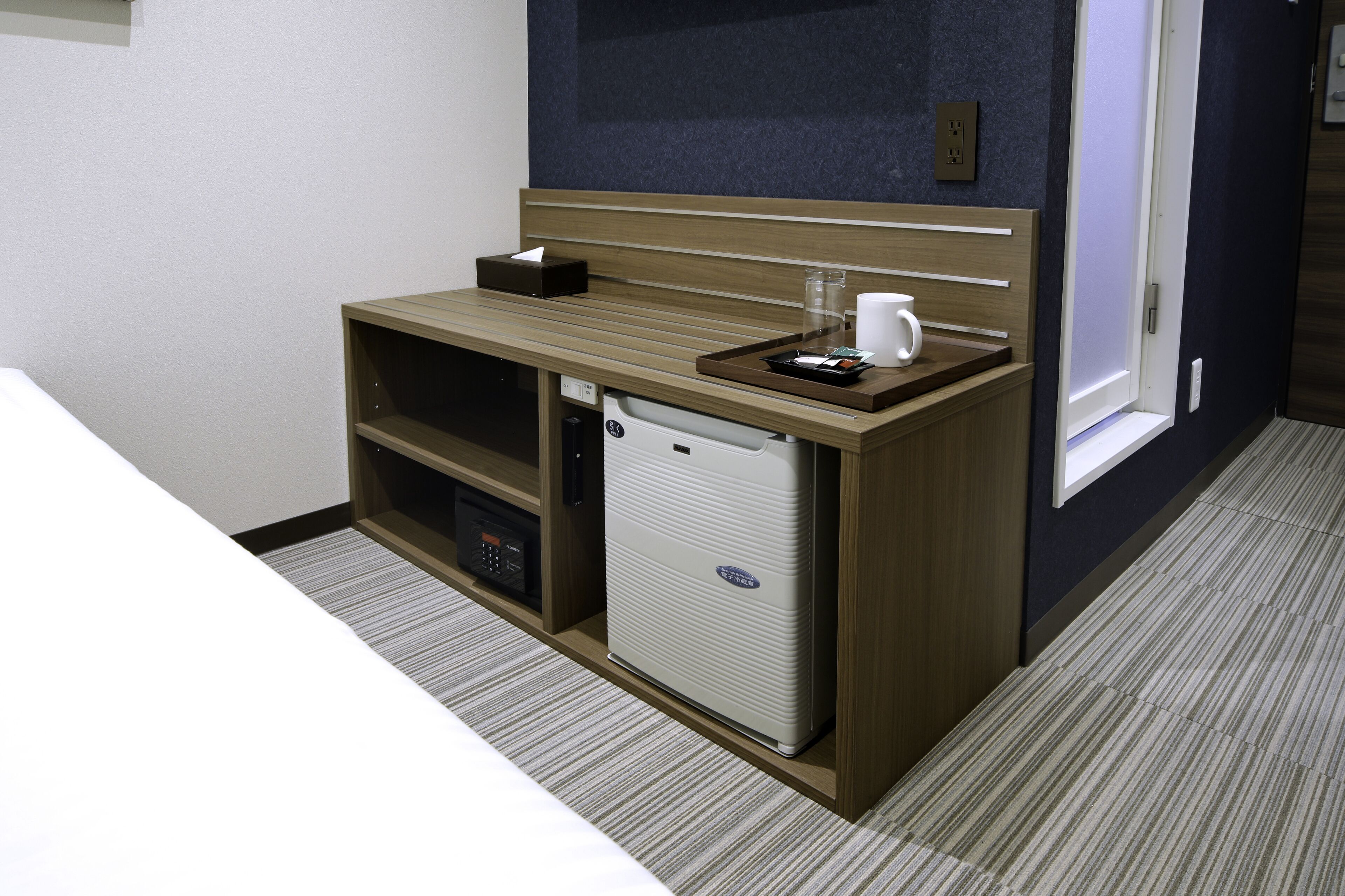 A hotel room with a compact minibar, a small refrigerator, a tray with a coffee cup, and a tissue box on a wooden counter.