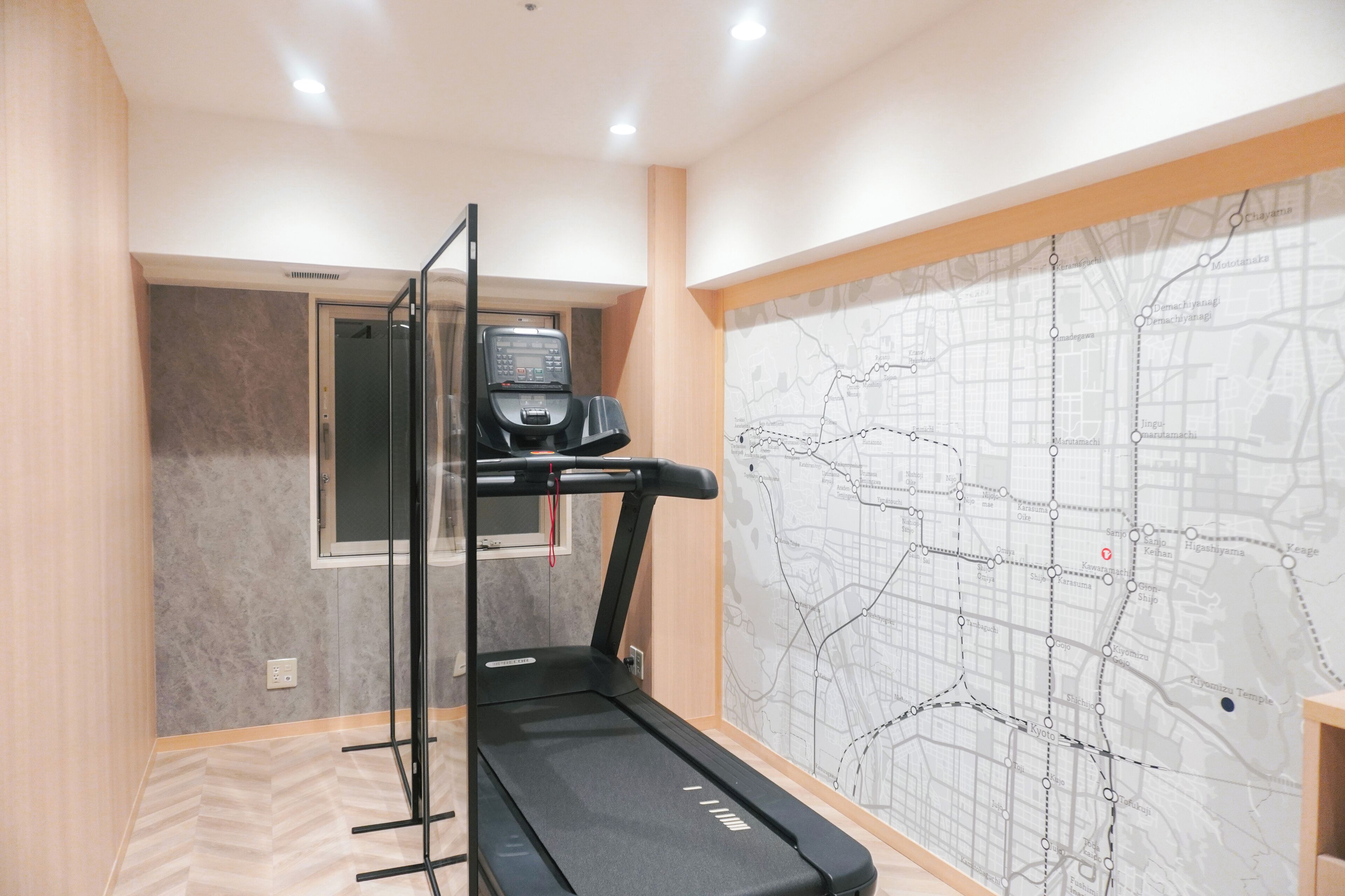 A hotel room with a treadmill, a map on the wall, and a window with a view of the city.