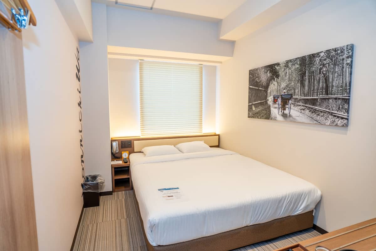 A hotel room with a large bed, a bedside table, a nightstand, a window with blinds, and a wall-mounted painting.