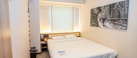 A hotel room with a large bed, a bedside table, a nightstand, a window with blinds, and a wall-mounted painting.