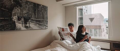 A bedroom with a bed, a window with a view of buildings, and a painting on the wall.